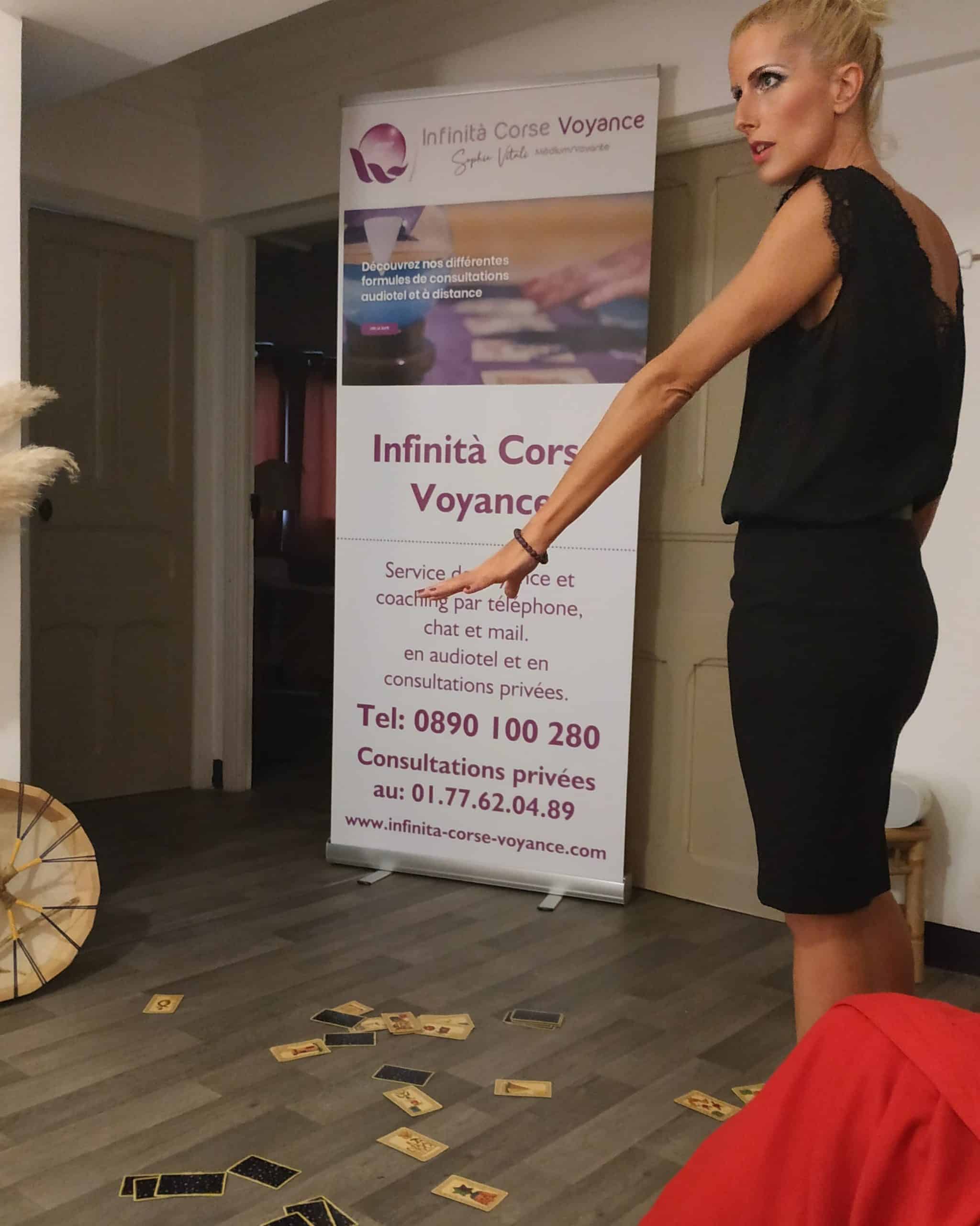 Sophie Vitali, célèbre médium, en pleine conférence pour Infinita Corse Voyance, expliquant un tirage de cartes avec l’Oracle de Belline, devant un kakemono promotionnel, cartes étalées au sol.